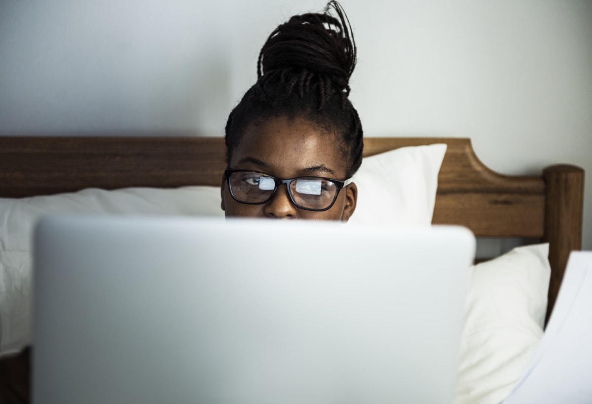 black woman wearing glasses sitting in bed looking at laptop screen