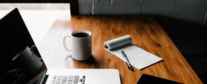 a wooden desk with laptop, phone, coffee cup, and notepad